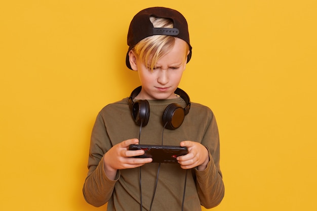 Young caucasian little boy using mobile phone against yellow, guy looks concentrated, holding his mobile phone