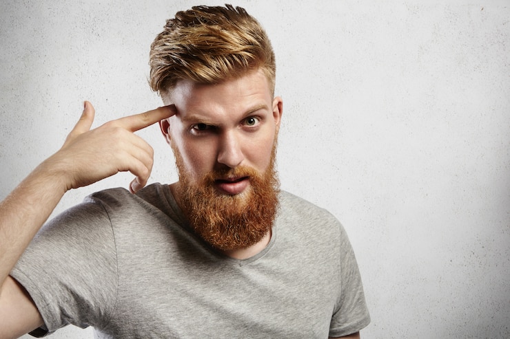 Free Photo young caucasian macho man with blond beard and hipster hair-style pointing finger at his temple meaning “use your brain!”. handsome guy in grey casual t-shirt is surprised and slightly irritated.