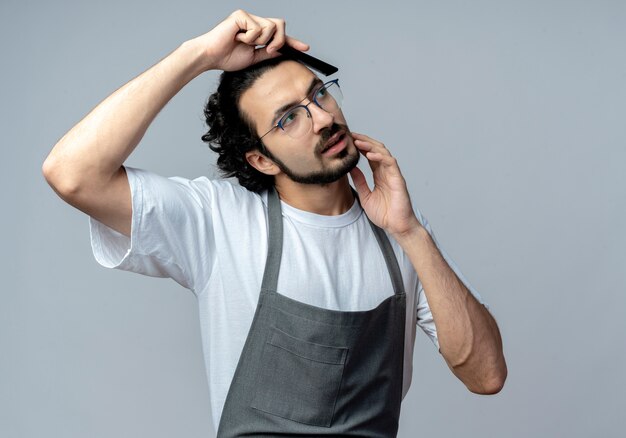 Young caucasian male barber wearing glasses and wavy hair band in uniform combing hair touching chin and looking at side isolated on white background with copy space