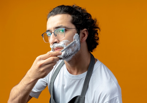 Young caucasian male barber wearing glasses and wavy hair band in uniform looking and shaving his own beard with straight razor with shaving cream put on his face