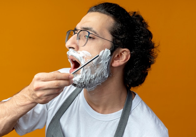 Young caucasian male barber wearing glasses and wavy hair band in uniform shaving his own beard with straight razor with shaving cream put on his face looking straight with open mouth