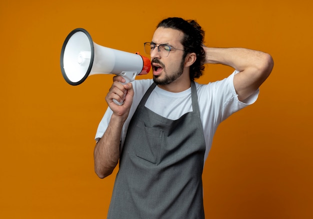 Young caucasian male barber wearing uniform and glasses shouting in loud speaker with closed eyes and hand behind head isolated on orange background