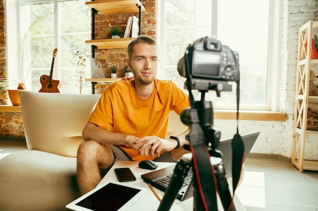 Young caucasian male blogger with professional camera recording video review of gadgets at home. Blogging, videoblog, vlogging. Man making vlog or live stream about photo or technical novelty.