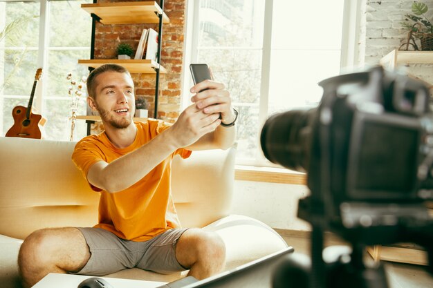 Young caucasian male blogger with professional camera recording video review of smartphone at home