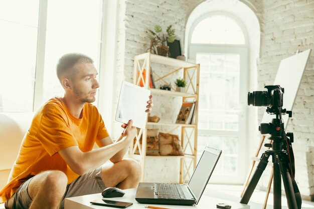 自宅でタブレットのビデオレビューを記録するプロのカメラを持つ若い白人男性ブロガー