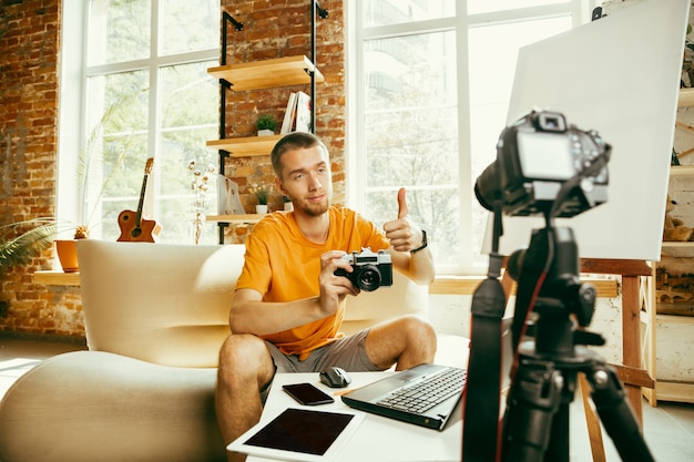Young caucasian male blogger with professional equipment recording video review of camera at home. Blogging, videoblog, vlogging. Man making vlog or live stream about photo or technical novelty.