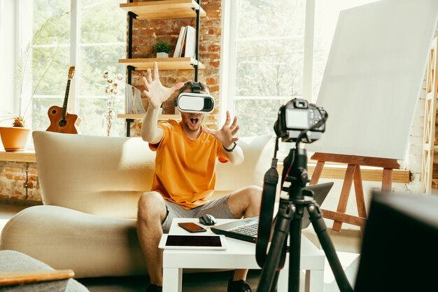 Young caucasian male blogger with professional equipment recording video review of VR glasses at home. Blogging, videoblog, vlogging. Man using virtual reality headset while streaming live.