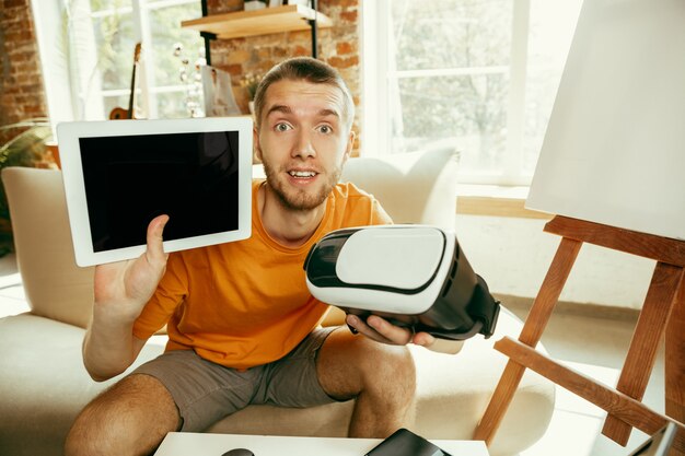 Young caucasian male blogger with professional equipment recording video review of VR glasses at home. Videoblog, vlogging. Man showing tablet and headset of virtual reality while streaming live.