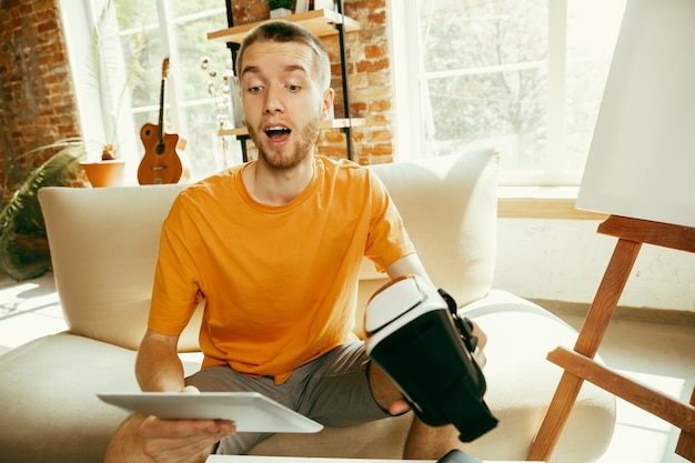 Young caucasian male blogger with professional equipment recording video review of VR glasses at home