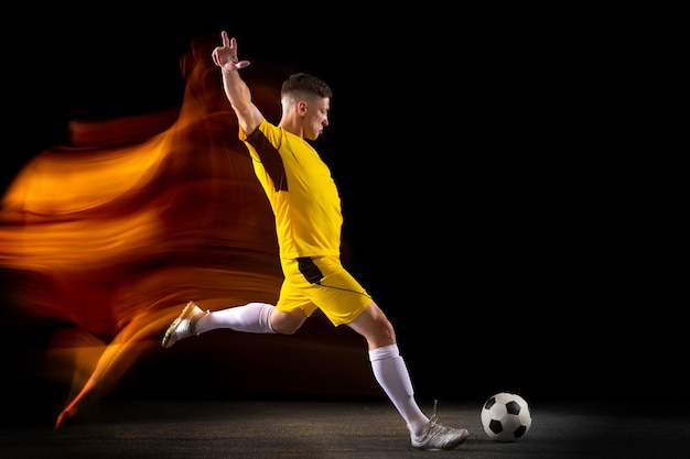 Young caucasian male football or soccer player kicking ball for the goal in mixed light on dark wall concept of healthy lifestyle professional sport hobby