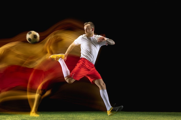 Young caucasian male football or soccer player in sportwear and boots kicking ball for the goal in mixed light on dark wall. Concept of healthy lifestyle, professional sport, hobby.