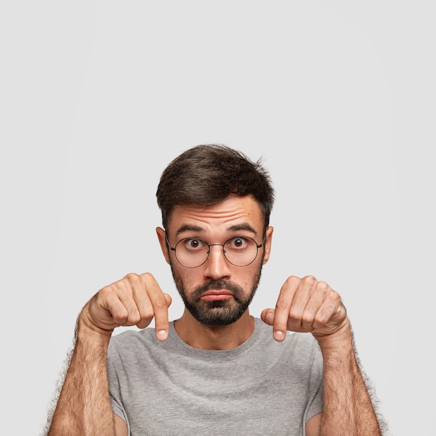 Young Caucasian male has surpised expression, points down with both index fingers, purses lips in bewilderment, demonstrates something, isolated over white wall with copy space upwards