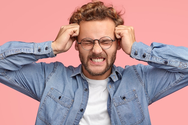 Foto gratuita il giovane maschio caucasico soffre di mal di testa ed emicrania, tiene i pugni sulle tempie, stringe i denti, ha un'espressione frustrata, vestito con una camicia alla moda in denim, isolato su un muro rosa. ragazzo malato