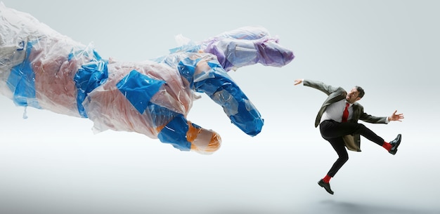 Young caucasian man avoiding big plastic hand on white  wall. Male model in office attire fighting in motion and action. Looks angry, scared, pressured. Eco problems, pollution, recycling.