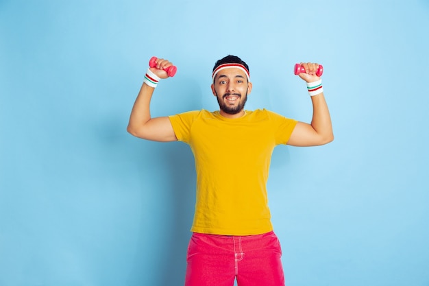 Giovane uomo caucasico in abiti luminosi, formazione su sfondo blu concetto di sport, emozioni umane, espressione facciale, stile di vita sano, gioventù, vendite. allenarsi con i pesi colorati. copyspace.