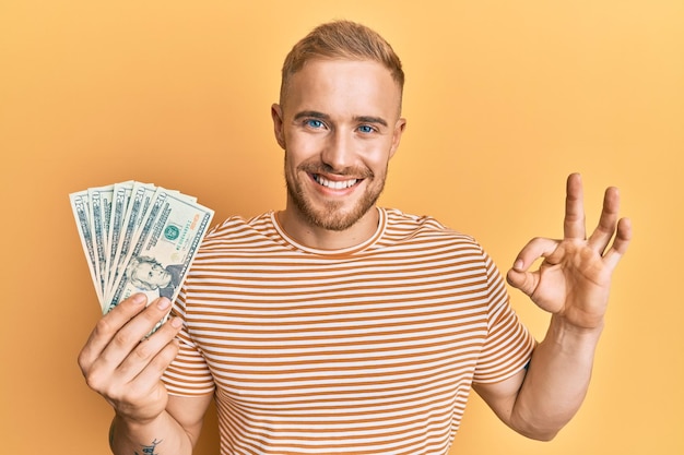 Young caucasian man holding dollars doing ok sign with fingers smiling friendly gesturing excellent symbol