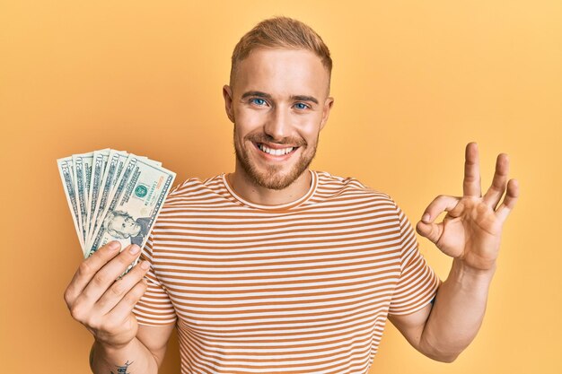 Young caucasian man holding dollars doing ok sign with fingers smiling friendly gesturing excellent symbol