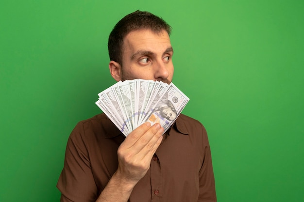 Young caucasian man holding money looking at side from behind it isolated on green background with copy space