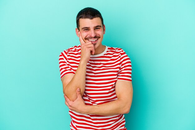 Young caucasian man isolated on blue background smiling happy and confident, touching chin with hand.