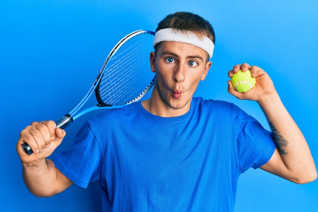 Free photo young caucasian man playing tennis holding racket and ball making fish face with mouth and squinting eyes, crazy and comical.