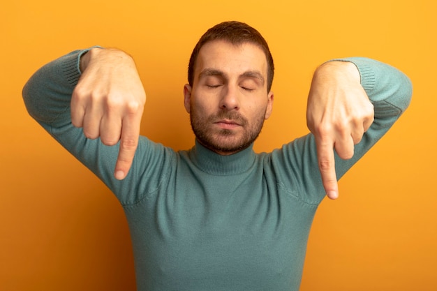 Free photo young caucasian man pointing down with closed eyes isolated on orange wall