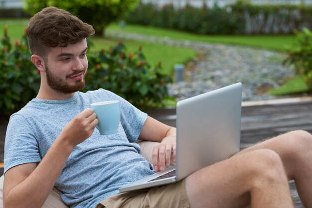 マグカップとラップトップの庭で横たわっている若い白人男