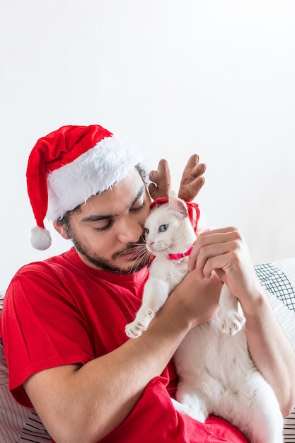 Молодой кавказец в шляпе Санта-Клауса играет со своим белым котом в лосиных рогах