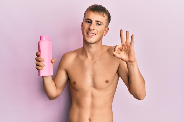 Free photo young caucasian man shirtless holding shampoo bottle doing ok sign with fingers smiling friendly gesturing excellent symbol