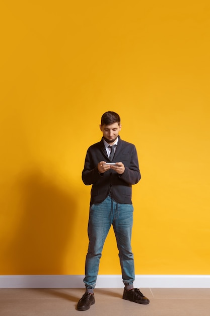 Free photo young caucasian man using smartphone full body length portrait isolated over yellow wall