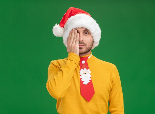 Young caucasian man wearing christmas hat and tie covering half of face with hand looking at camera isolated on green background with copy space