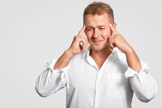 Young Caucasian man with concntrated facial expression keeps fore fingers on temples, focused down, thinks about something, tries to remember necessary information, isolated on white wall.
