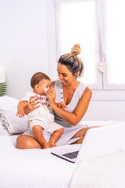 Young caucasian mother with her son in the room on the bed teleworking and caring for her child