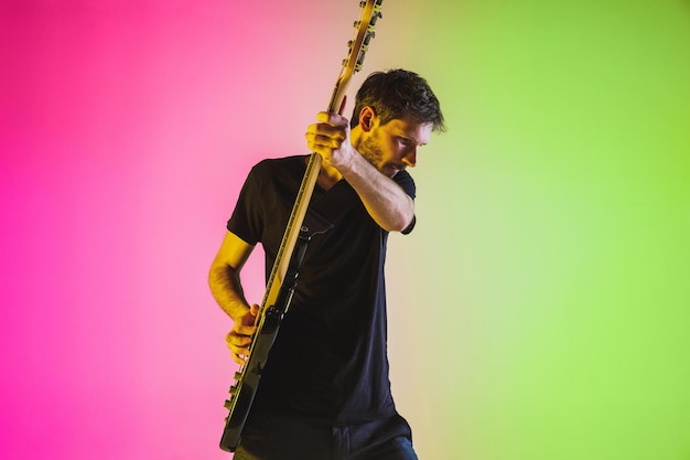 Free photo young caucasian musician playing bass guitar in neon light on pink-green