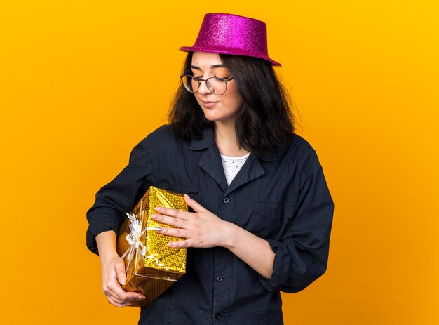 Young caucasian party girl wearing party hat and glasses holding and looking at gift package isolated on orange wall