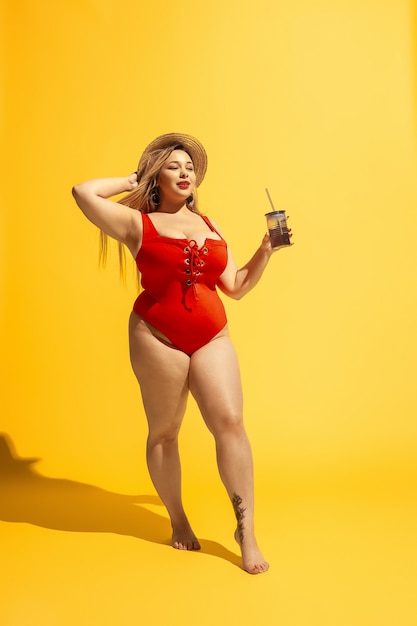 Young caucasian plus size female model's preparing for beach resort on yellow wall. Woman in red swimsuit and hat drinking cocktail. Concept of summertime, party, body positive, equality.