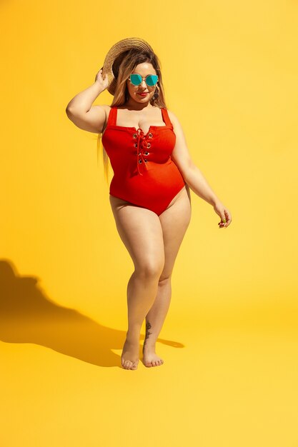 若い白人のプラスサイズの女性モデルが黄色い壁のビーチリゾートの準備をしています。赤い水着、帽子、サングラスの女性。夏のコンセプト、パーティー、ボディポジティブ、平等、そして寒さ。