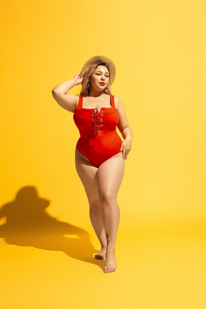 Young caucasian plus size female model's preparing vacations on yellow wall. Woman in red swimsuit and hat posing confident.