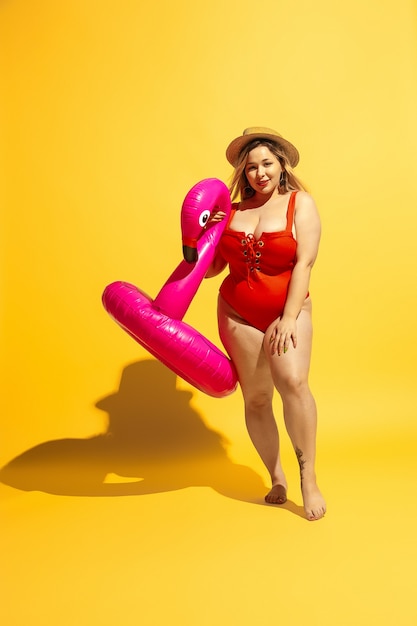 Young caucasian plus size female model's preparing vacations on yellow wall. Woman in red swimsuit and hat posing with swimring.