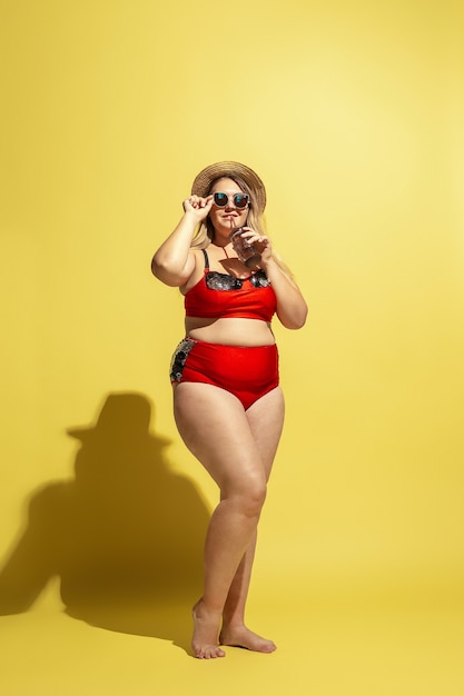 若い白人のプラスサイズの女性モデルが、黄色い壁に休暇の準備をしている。赤い水着、帽子、サングラスの女性。