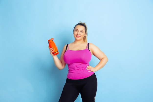 Young caucasian plus size female model's training on blue background. Concept of sport, human emotions, expression, healthy lifestyle, body positive, equality. Posing with bottle, copyspace.