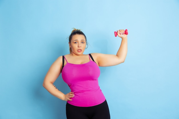 Free photo young caucasian plus size female model's training on blue background. concept of sport, human emotions, expression, healthy lifestyle, body positive, equality. training with the weights, copyspace.