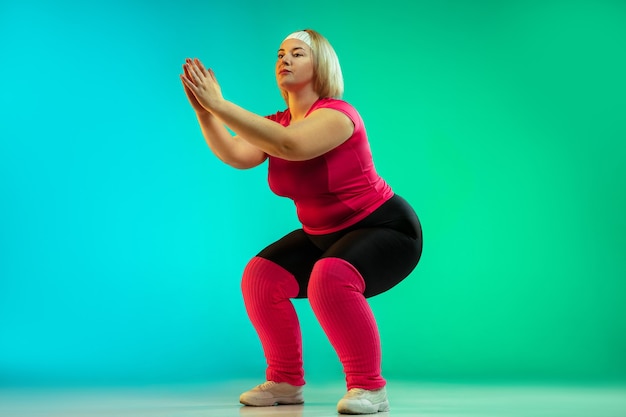 Young caucasian plus size female model's training on gradient green background in neon light. Doing workout exercises, stretching, cardio. Concept of sport, healthy lifestyle, body positive, equality.