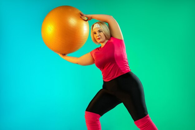 네온 불빛에 그라데이션 녹색 배경에 젊은 백인 플러스 크기 여성 모델의 교육. fitball로 운동 운동하기. 스포츠, 건강한 라이프 스타일, 신체 긍정적, 평등의 개념.