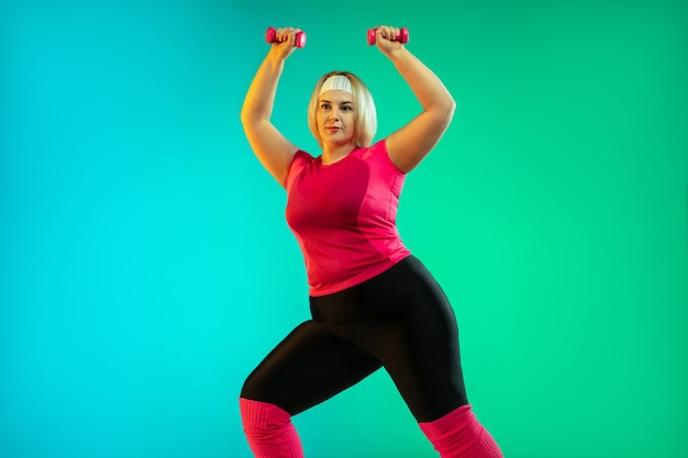 Young caucasian plus size female model's training on gradient green background in neon light. Doing workout exercises with the weights. Concept of sport, healthy lifestyle, body positive, equality.