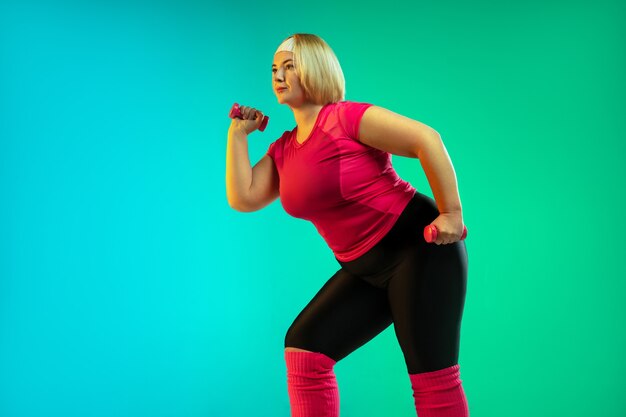 Young caucasian plus size female model's training on gradient green background in neon light. Doing workout exercises with the weights. Concept of sport, healthy lifestyle, body positive, equality.