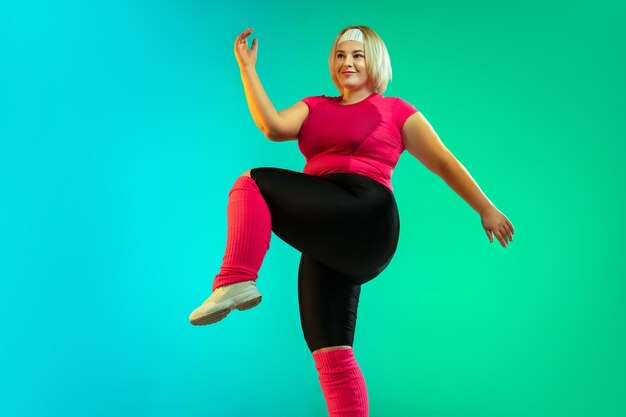 Young caucasian plus size female model's training on gradient green background in neon light.