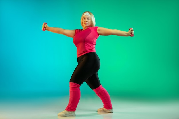 Young caucasian plus size female model's training on gradient green background in neon light.