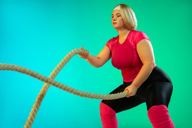 Young caucasian plus size female model's training on gradient green background in neon light.