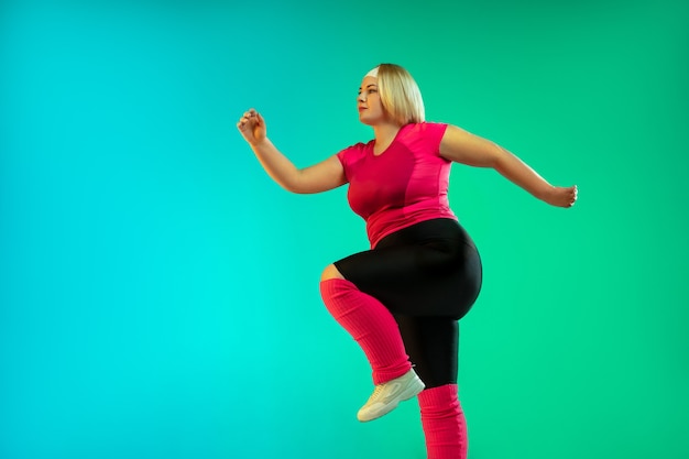 Young caucasian plus size female model's training on gradient green background in neon light.