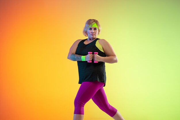 Young caucasian plus size female model's training on gradient orange background in neon light. Doing workout exercises with the weights. Concept of sport, healthy lifestyle, body positive, equality.